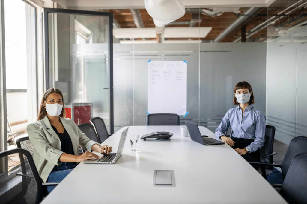 Two women wearing masks in a conference room with laptops in front of them.