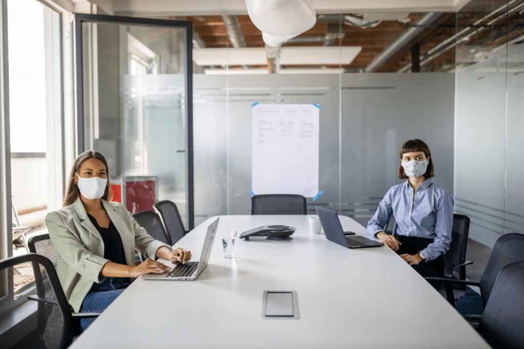 Dos mujeres enmascaradas en una sala de conferencias con ordenadores portátiles delante.